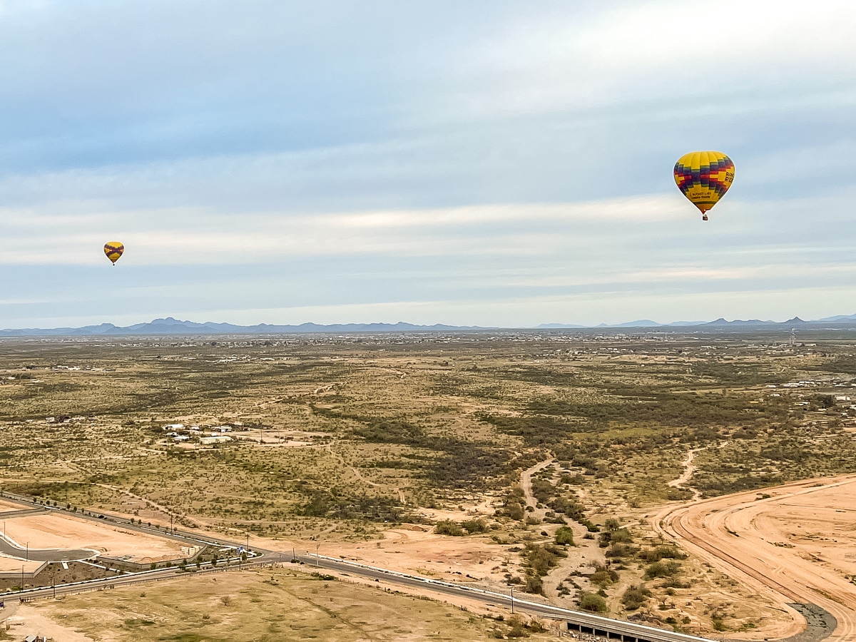 Two hot air balloons float over a vast, arid landscape with sparse vegetation, mountains in the distance, and roads crossing the foreground.