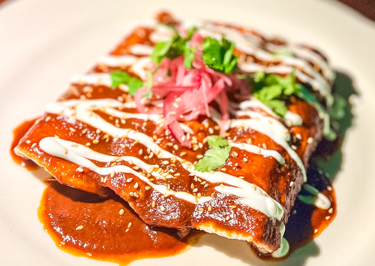 A plate of enchiladas garnished with cilantro, pickled onions, and drizzled with white sauce. The enchiladas are covered in a red sauce and sprinkled with sesame seeds.