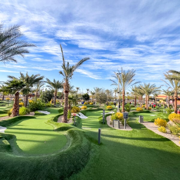 A well-maintained miniature golf course with lush green turf, palm trees, and various obstacles under a partly cloudy sky.