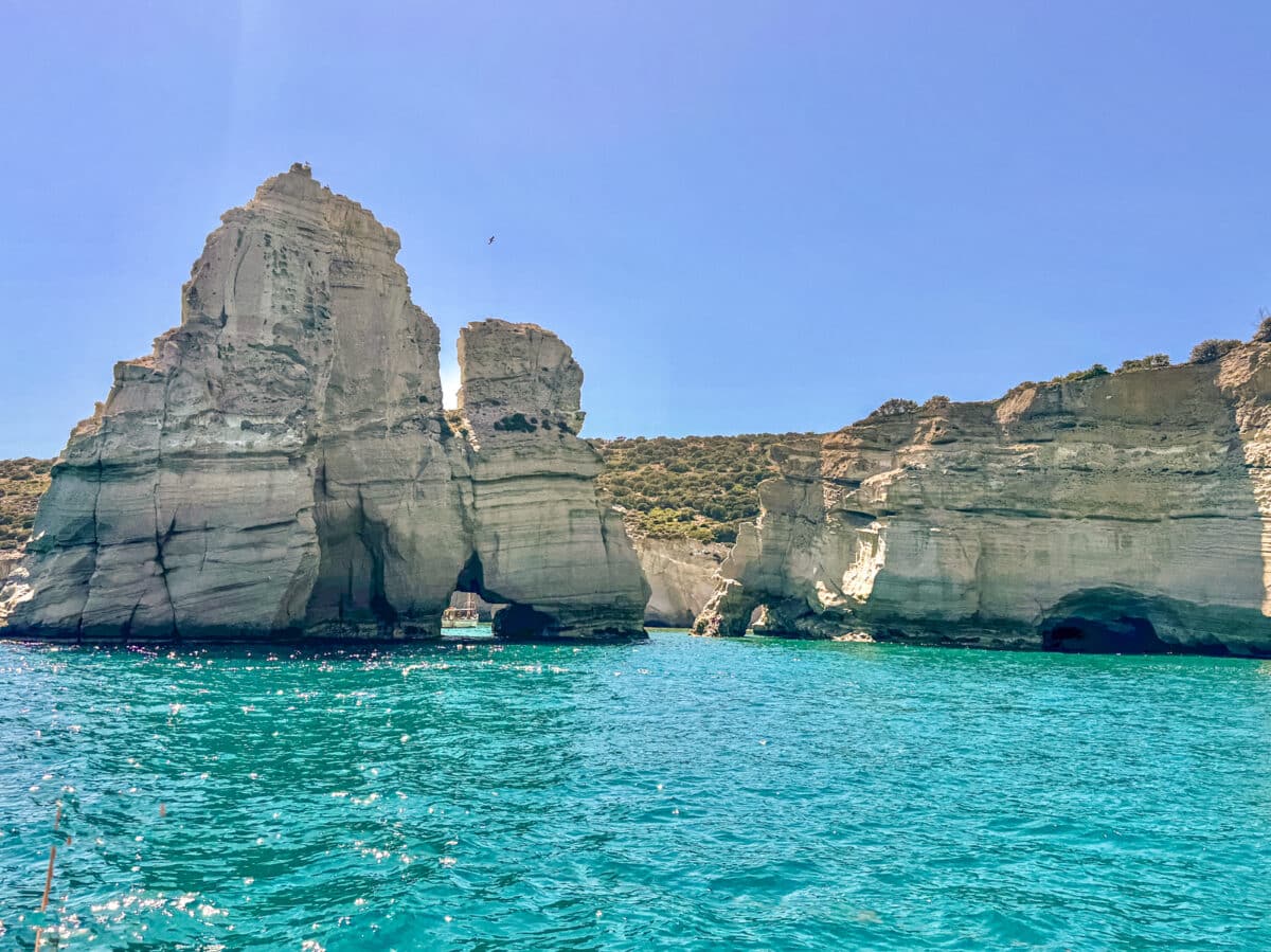 Rock formations rise from clear turquoise water under a bright blue sky. Several caves and arches punctuate the rocks, with one prominent arch near the center. Sparse vegetation covers the top, creating a stunning scene reminiscent of an Andrea Updyke portfolio.