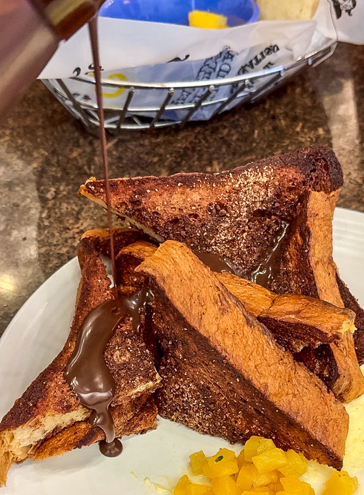 Thick chocolate syrup being poured onto a stack of toasted bread slices on a white plate, accompanied by a side of chopped fruits in the background.