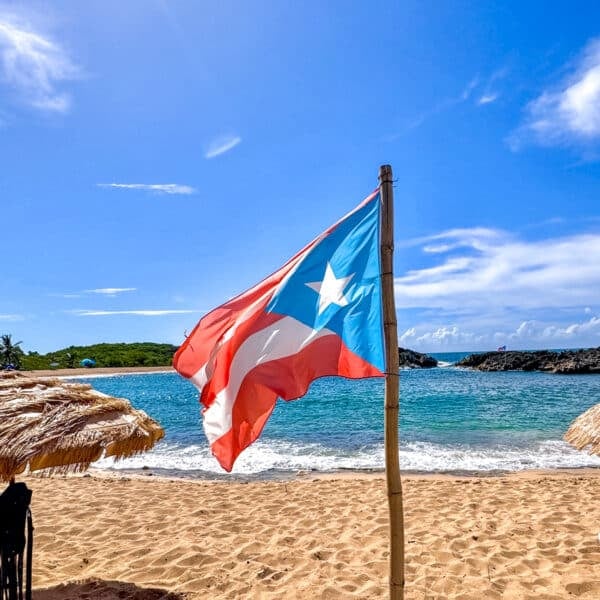 A Puerto Rican flag waves on a sunny beach with clear blue skies, straw umbrellas, and ocean waves in the background.