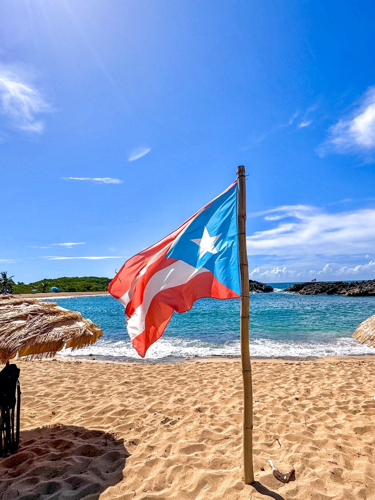 A Puerto Rican flag waves on a sunny beach with clear blue skies, straw umbrellas, and ocean waves in the background.