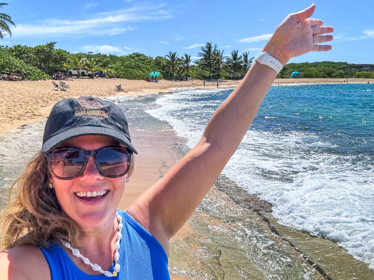 A woman wearing sunglasses and a cap smiles while raising her arm on a sandy beach with waves and palm trees in the background.