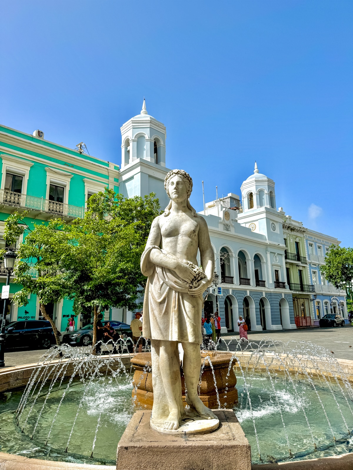 A statue of a woman holding flowers stands at the center of a fountain, surrounded by historic buildings with ornate architecture in a sunny plaza.