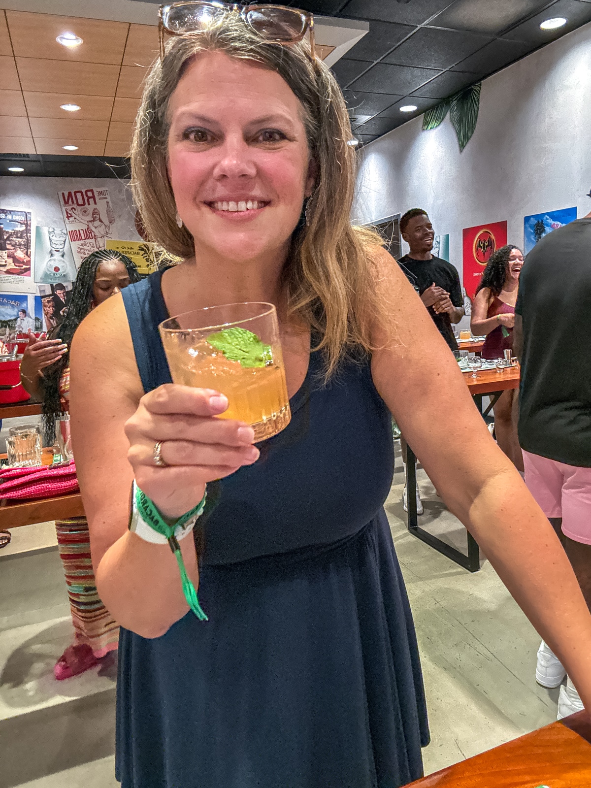 Woman in a navy blue dress holding a cocktail with mint garnish, standing in a lively indoor setting with other people in the background.