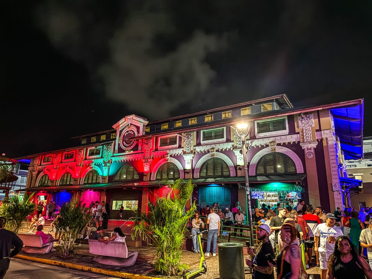 A crowd of people stands and sits in front of a large, colorfully lit building at night, with red, green, and blue lights illuminating its facade.