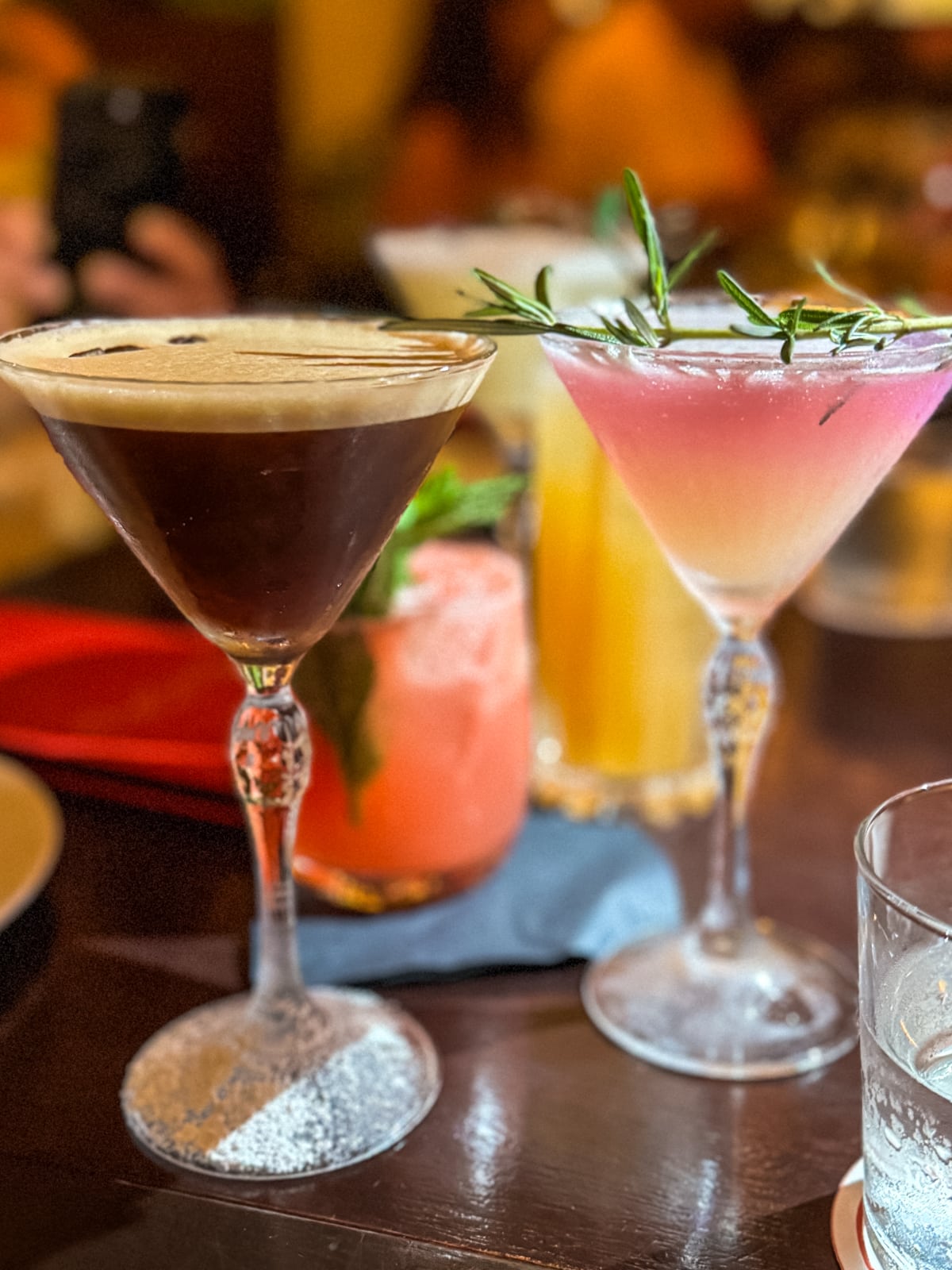 A variety of cocktails, including a dark one in a martini glass, a pink drink garnished with rosemary, a vibrant red drink, and a yellow beverage, are arranged on a table.