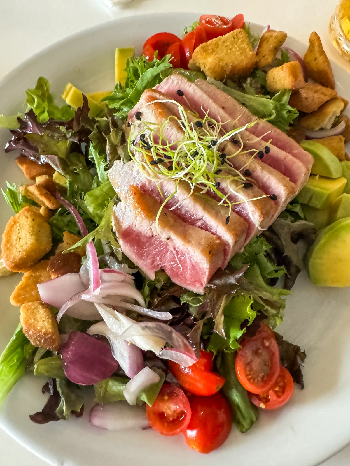 A plate of salad with seared tuna slices, tomatoes, onions, croutons, avocado, and leafy greens, garnished with microgreens.