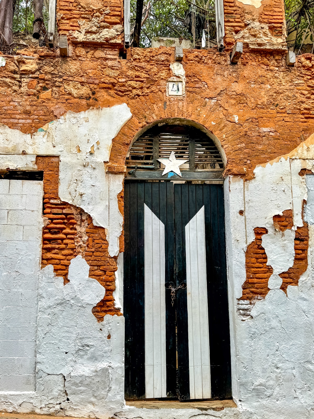 A tall wooden door with the Puerto Rican flag design is set in a weathered, partially crumbling brick wall with peeling white paint. The door is locked with a chain and padlock.