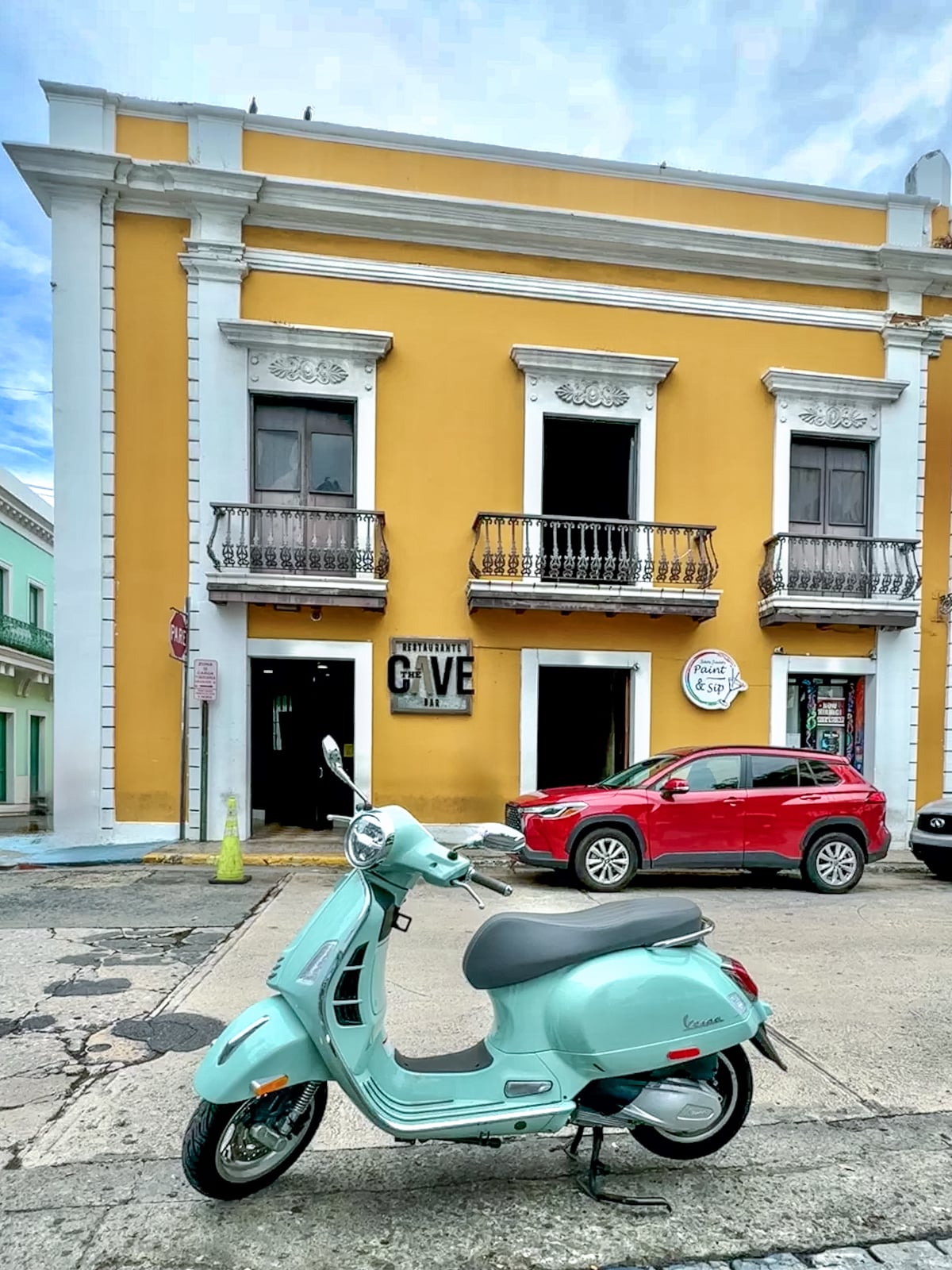 A light blue scooter is parked in front of a yellow two-story building with balconies on the second floor. A red car is parked on the side, and the building has signs for "Cave" and "Peace of Mind.