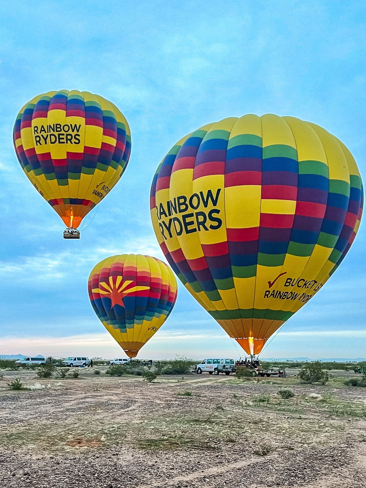 Three colorful hot air balloons with "Rainbow Ryders" labels are floating in the sky above a grassy area with several parked vehicles.