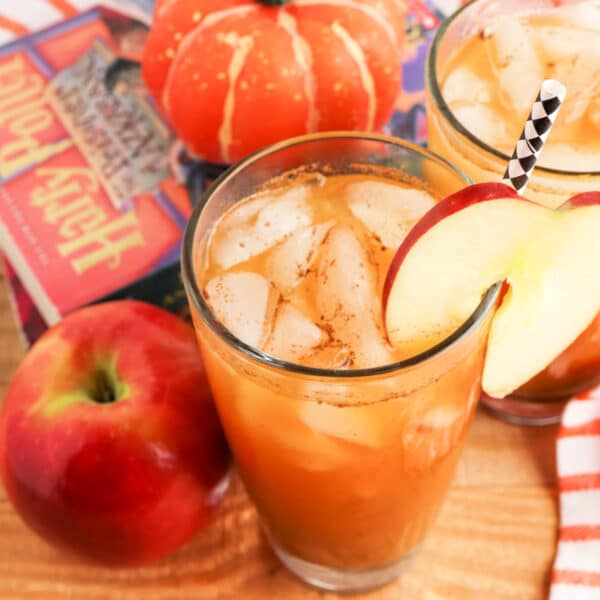 A glass filled with an iced beverage garnished with a slice of apple, next to a fresh apple, small pumpkin decor, and part of a colorful book cover.