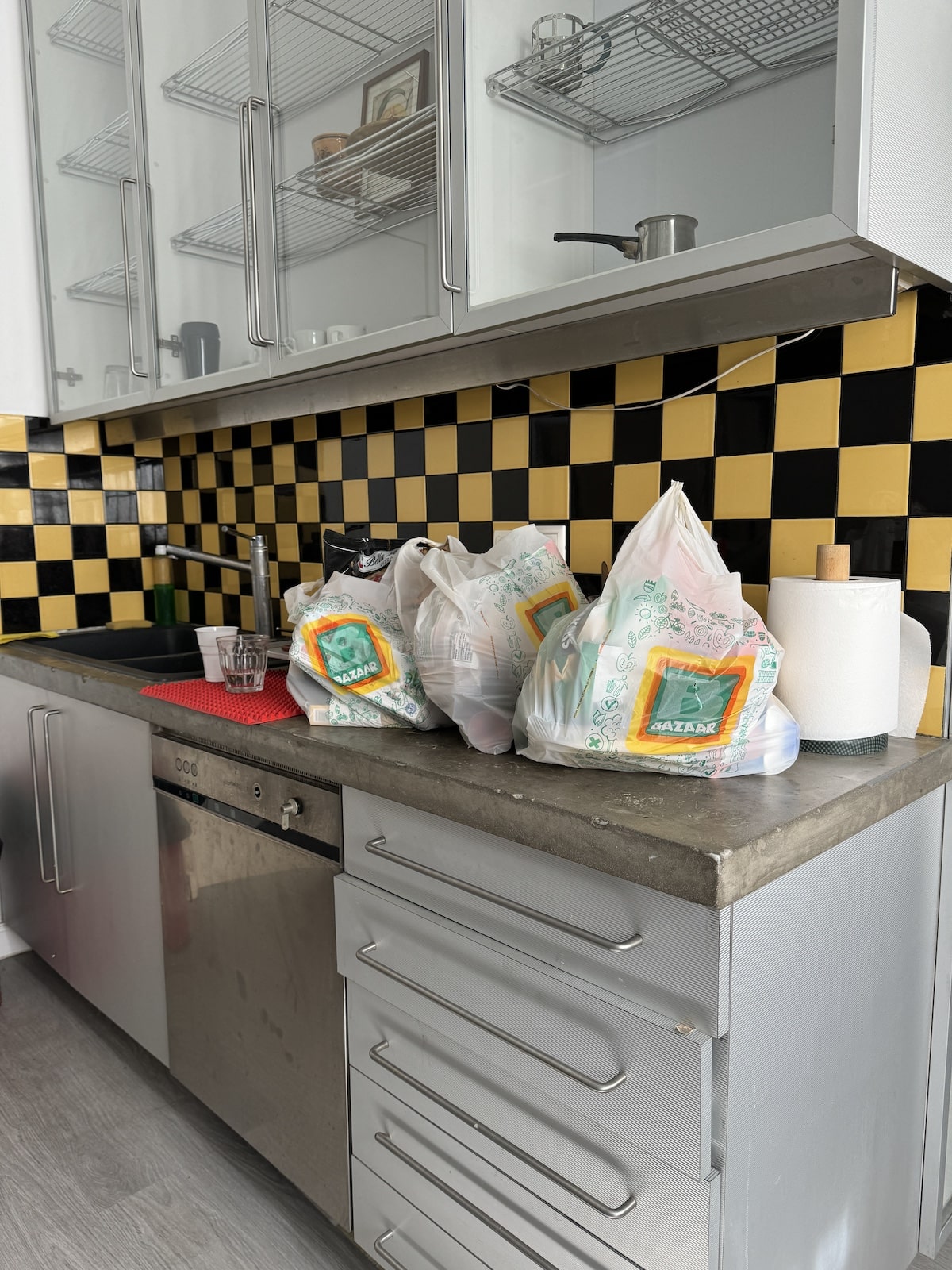 A kitchen counter with a concrete top has several grocery bags, a roll of paper towels, and a red drying mat. The backsplash features a yellow and black checkered pattern.