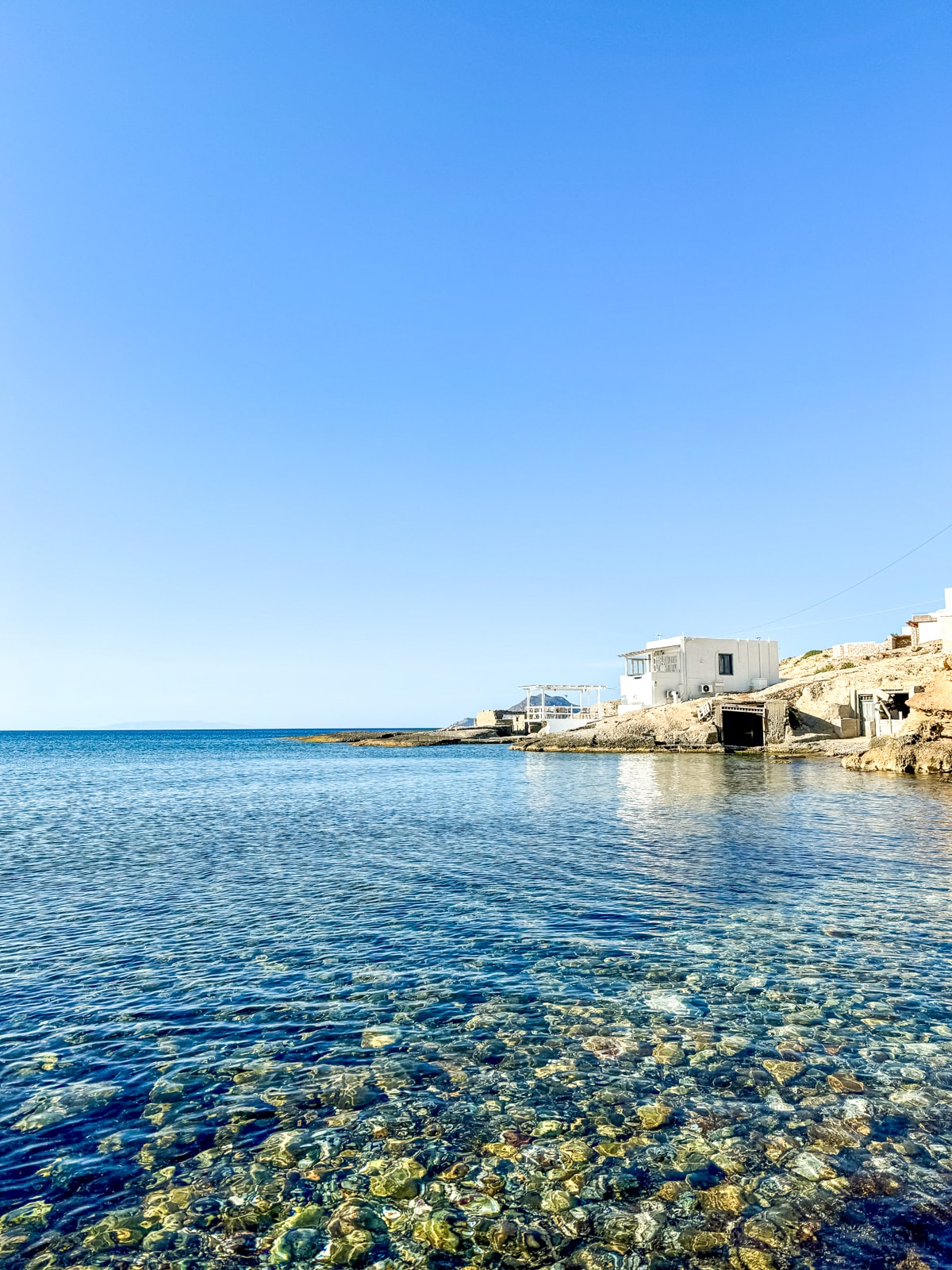 Clear, calm sea waters with visible rocks beneath, and a coastal village with white buildings in the background under a clear blue sky.