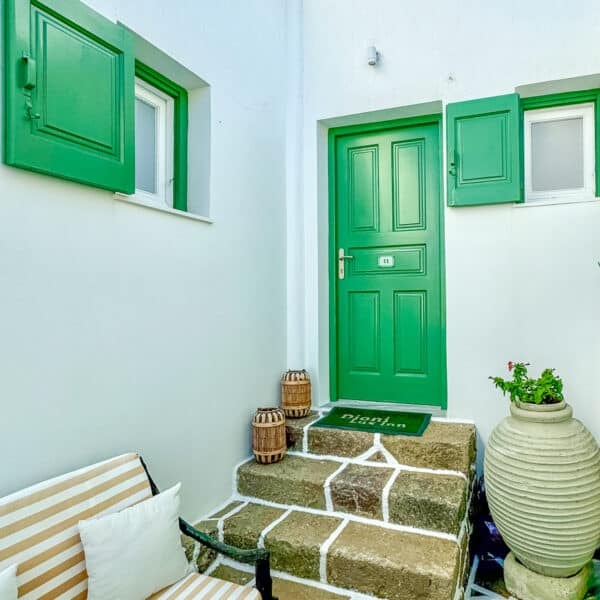 A green door with matching shutters, stone steps, potted plants, two barrels, and a striped bench cushion in a small courtyard.