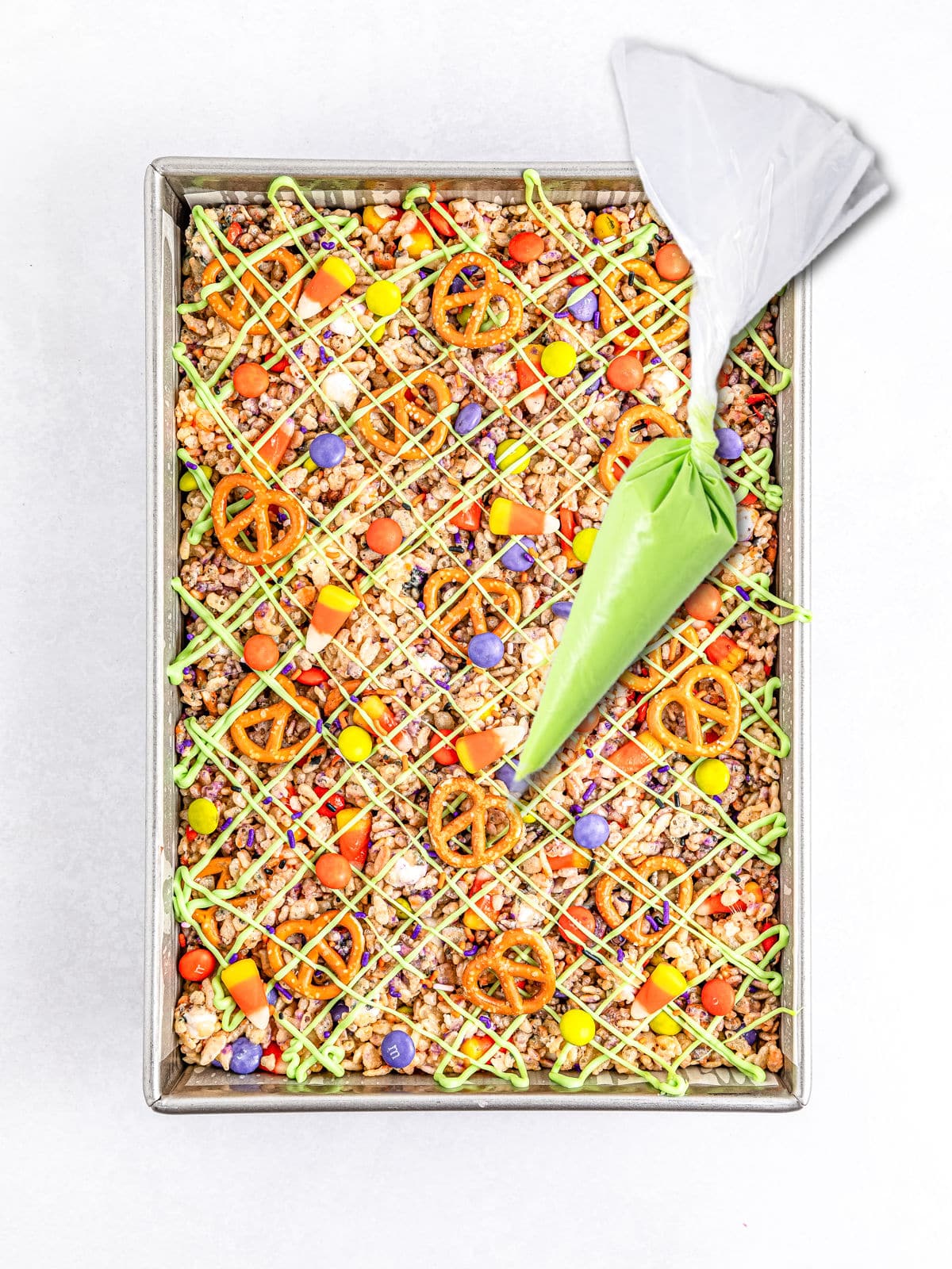 A rectangular tray containing a mixed snack of pretzels, colorful candies, and popcorn, drizzled with green icing, conjuring the spirit of Halloween rice krispie treats. Nearby, a piping bag filled with green icing is ready for use.