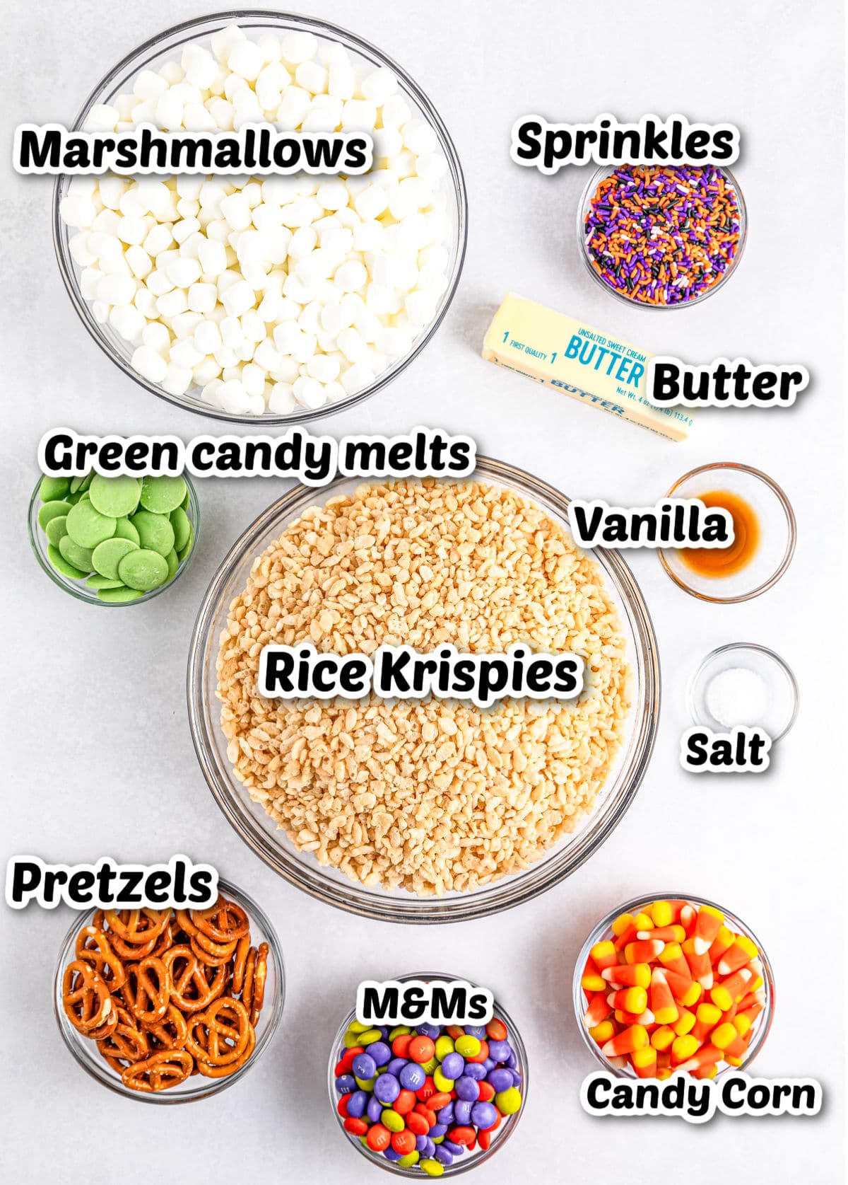 Ingredients for a Halloween Rice Krispies treats recipe: marshmallows, sprinkles, butter, vanilla, salt, green candy melts, rice krispies, pretzels, M&Ms, and candy corn arranged in bowls.