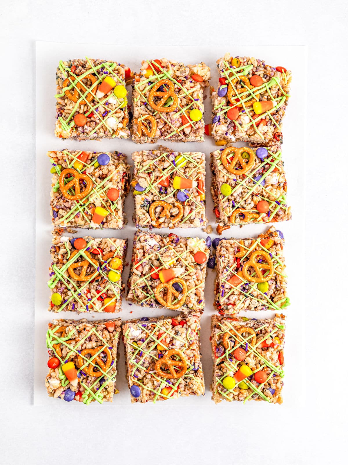 Nine colorful granola squares topped with pretzels, candy-coated chocolates, and two types of drizzle arranged on a white surface, reminiscent of festive Halloween Rice Krispie treats.