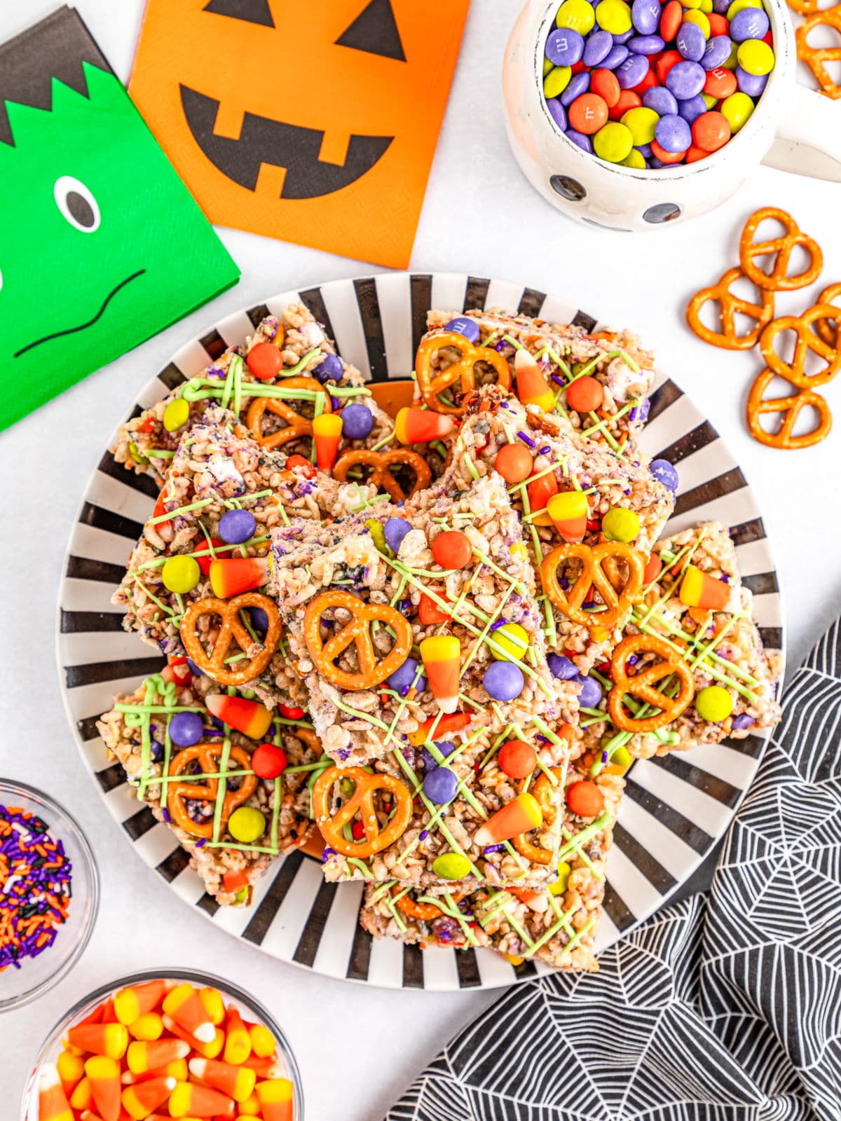 A plate of colorful Halloween Rice Krispie Treats topped with pretzels, candy corn, and drizzle, surrounded by bowls of candy and festive decorations.