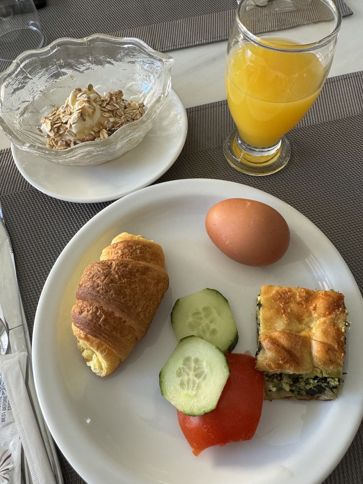 A breakfast plate with a croissant, cucumber slices, tomato, hard-boiled egg, and a spinach pastry. A bowl of yogurt with cereal and a glass of orange juice are beside the plate.