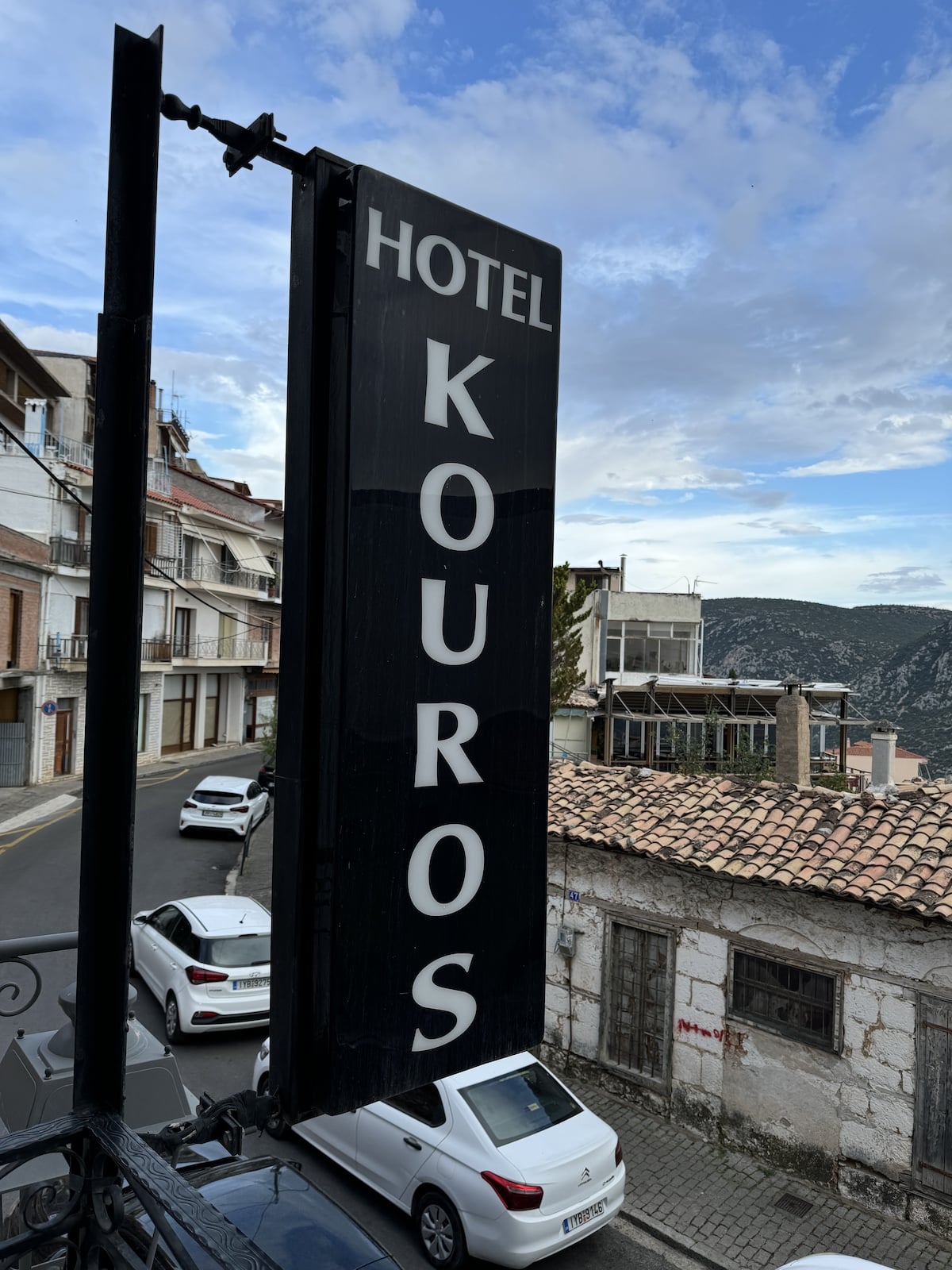 Vertical sign reading "Hotel Kouros" on a street with parked cars, old buildings, and a view of distant hills under a partly cloudy sky.