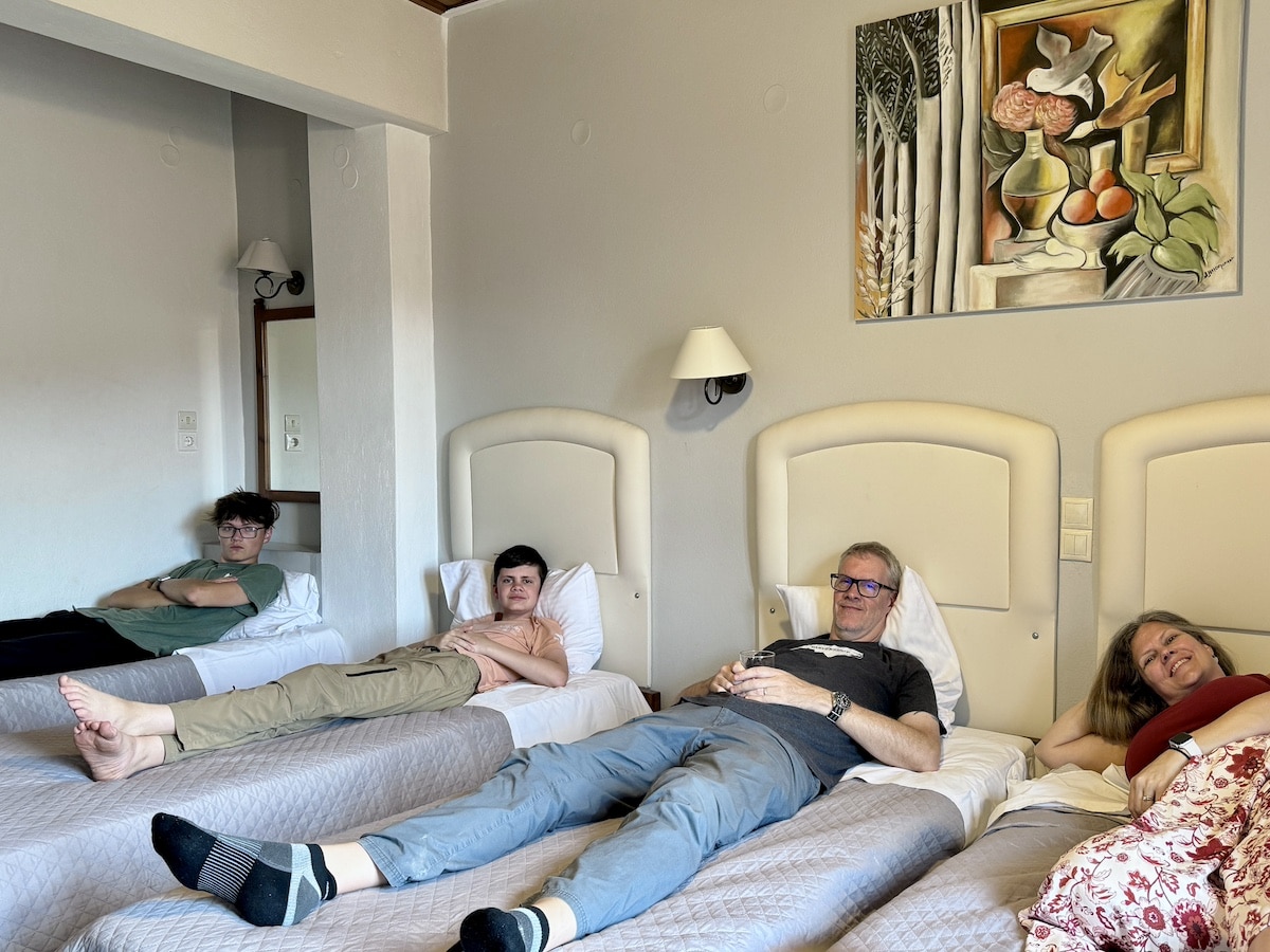 Four people are laying on separate beds in a hotel room, looking relaxed. The room has neutral-colored decor, a modern painting on the wall, and a lamp between two of the beds.