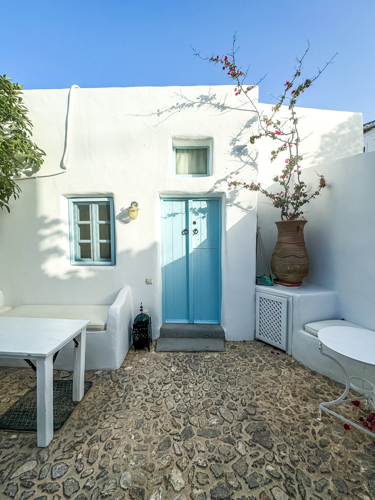 A quaint outdoor space features a stone floor, white walls, a light blue door, a potted plant climbing the wall, and white furniture, including a table with an attached bench and a small round table.