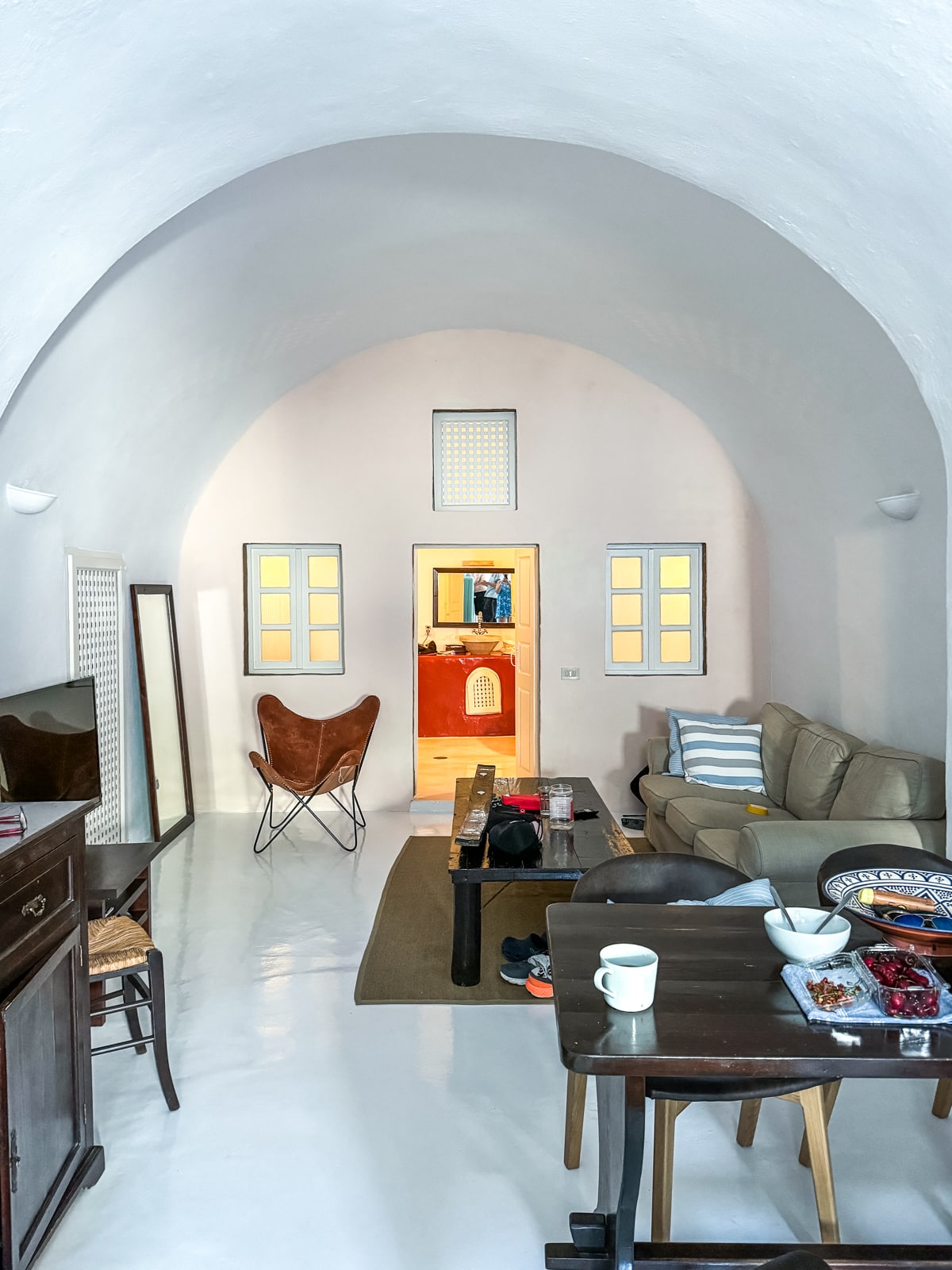 A tidy, well-lit living room with arched ceilings, a beige couch, a wooden coffee table, and a butterfly chair. A small dining area with a table and chairs is in the foreground.