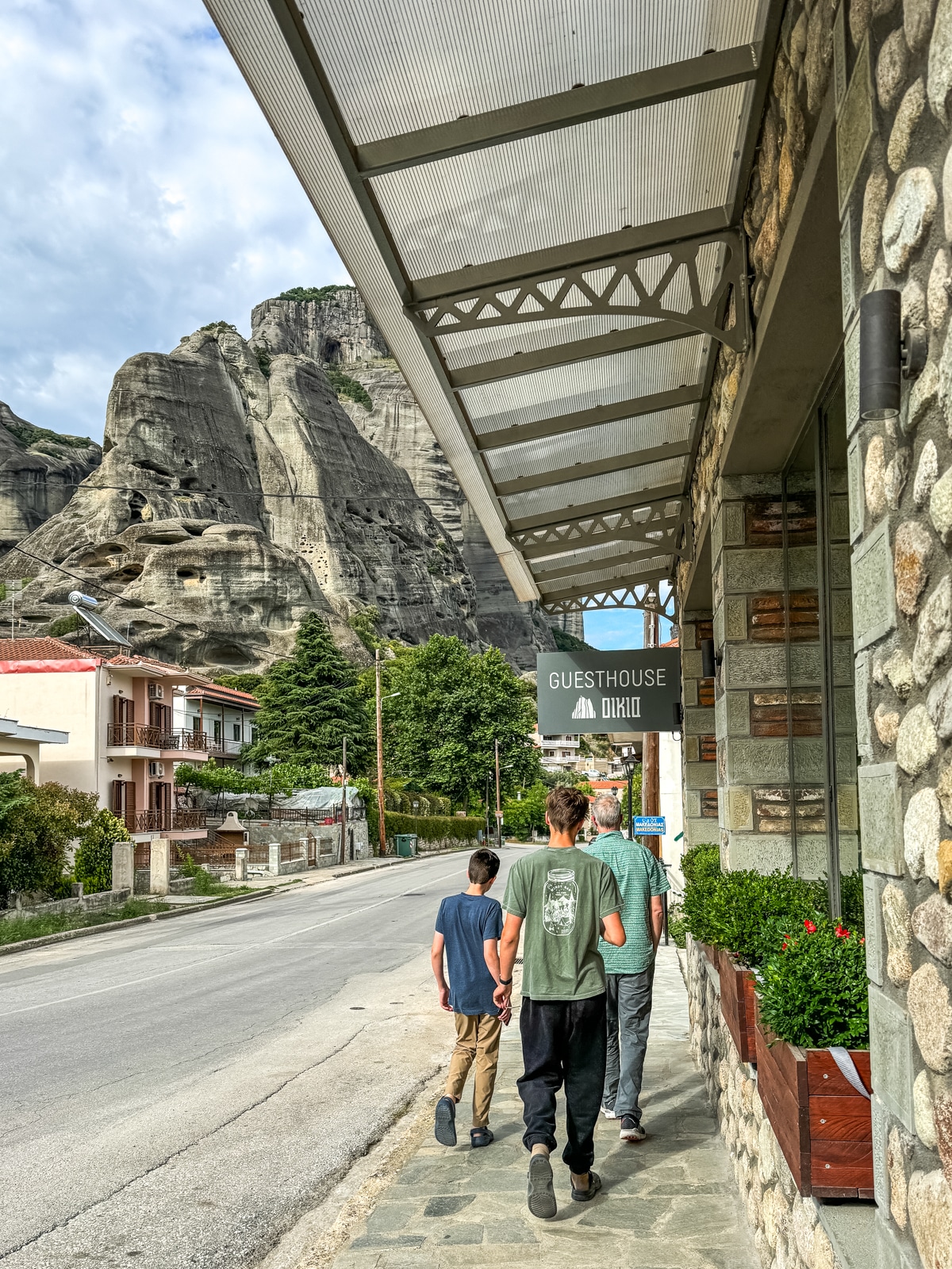 Three people walk down a sidewalk beside a stone-sided building with a "Guesthouse" sign. Rocky cliffs and residential buildings are seen in the background under a cloudy sky.