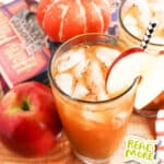 A glass of iced pumpkin juice garnished with an apple slice, with a red apple, pumpkins, and a book in the background. The text reads "Just is a Four Letter Word" at the top and "Pumpkin Juice" at the bottom.