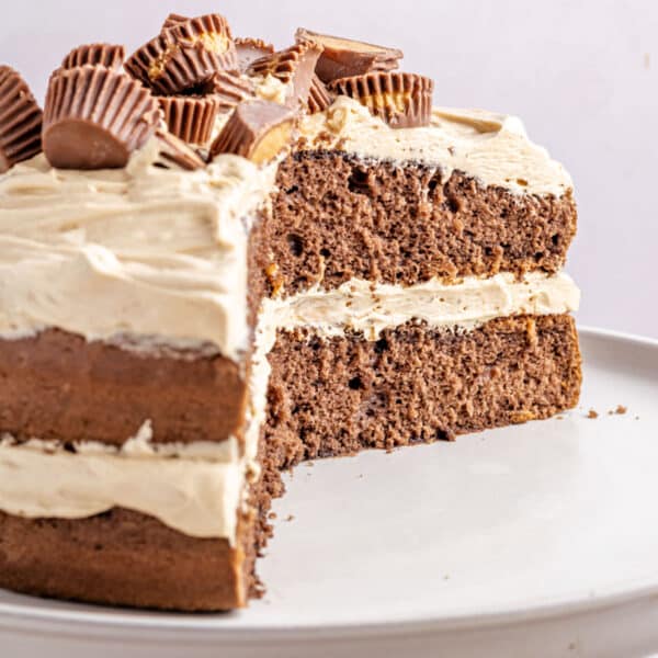 A chocolate layer cake with beige frosting, topped with mini peanut butter cups, partially sliced on a white plate.