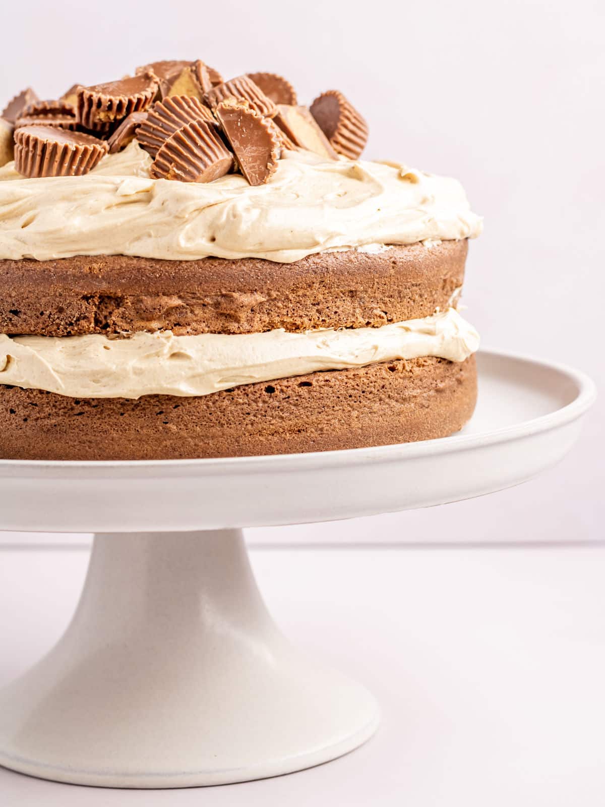 A chocolate cake with creamy peanut butter frosting and topped with pieces of peanut butter cups sits on a white cake stand.