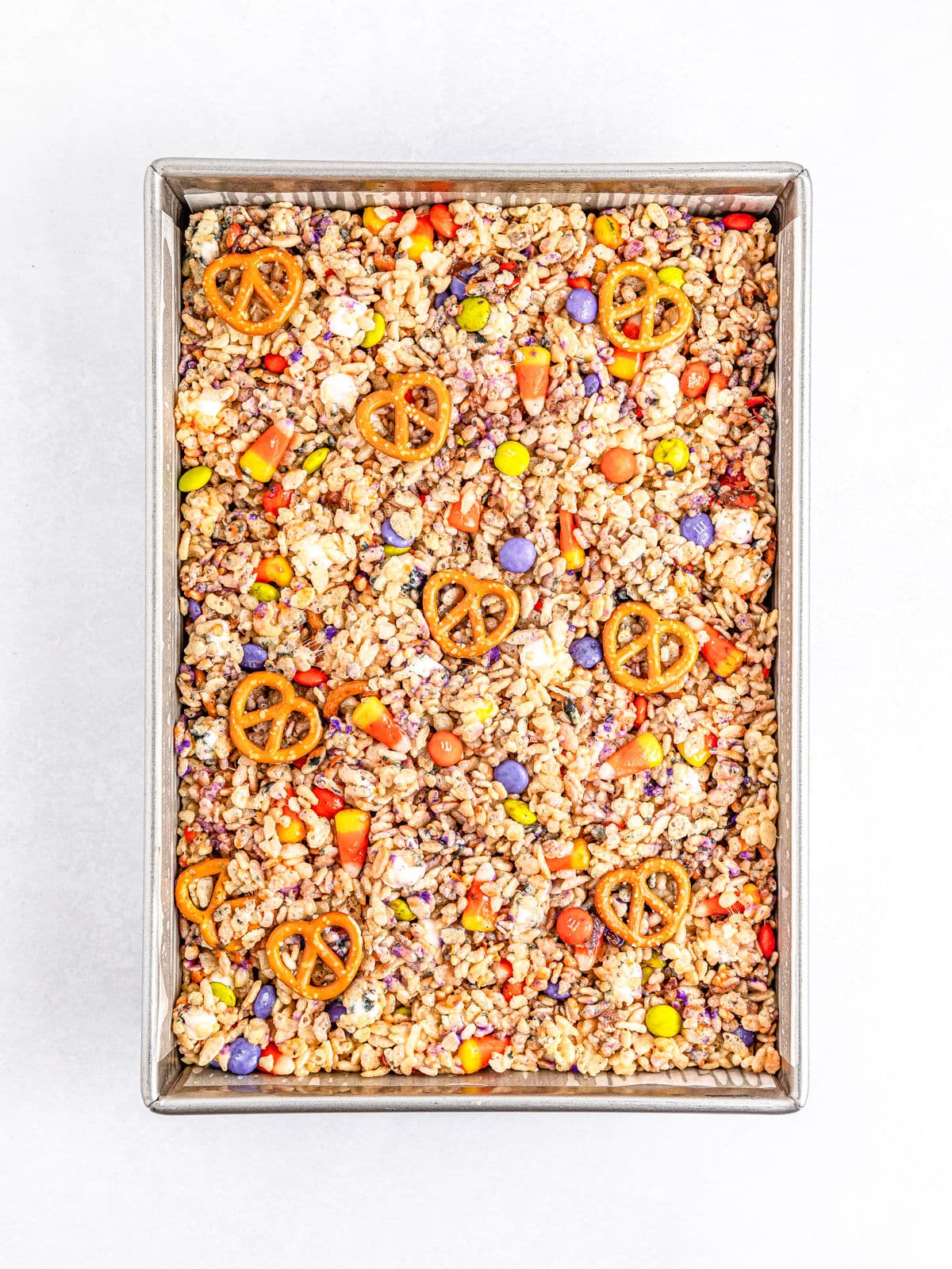 Rectangular baking tray filled with a colorful mix of pretzels, chocolate candies, and cereal, forming a snack mix on a white background, perfect for making Halloween Rice Krispie treats.