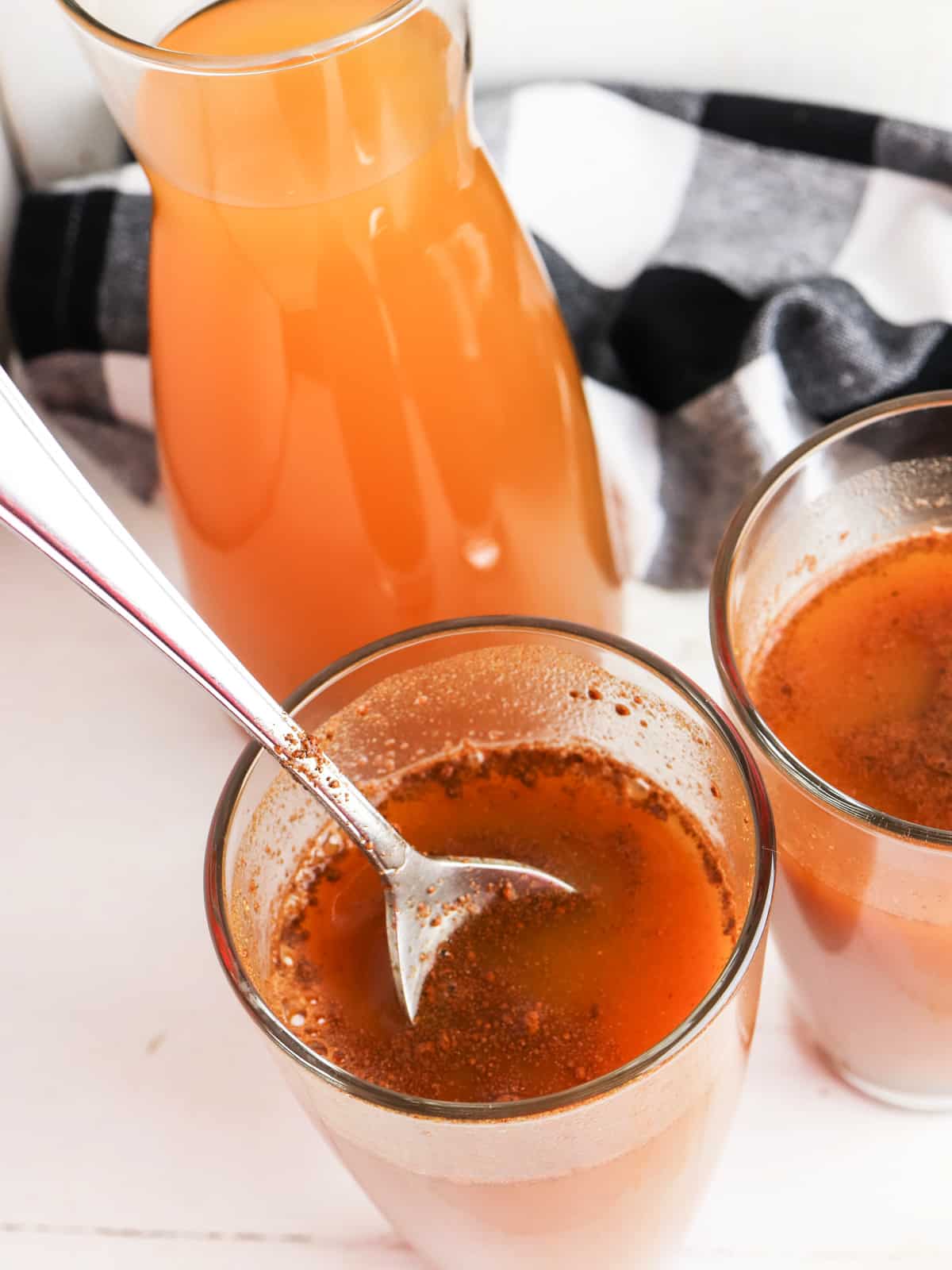 A glass pitcher and two glasses filled with an orange liquid, with one glass containing a spoon. A black and white cloth is in the background.