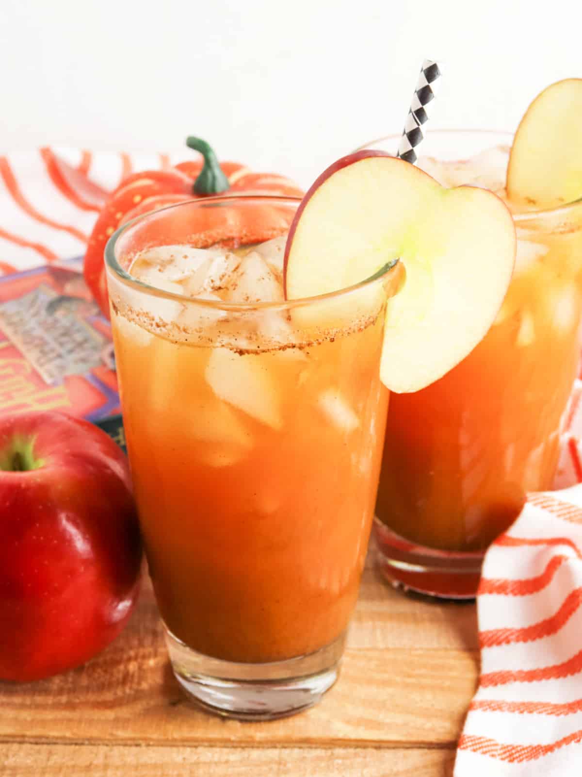 Two glasses of iced apple cider garnished with apple slices and paper straws. An apple and an orange-striped napkin are nearby.