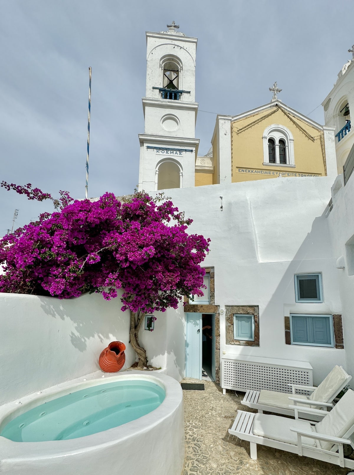 A small courtyard with a white plaster building, vibrant bougainvillea, a hot tub, and benches. A bell tower and the roofline of a church are visible in the background.