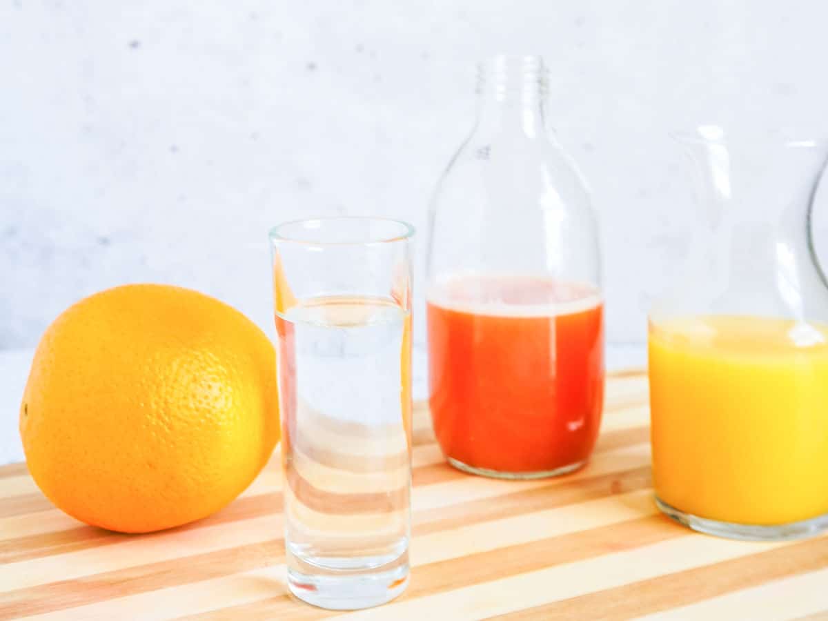 Glass of water, whole orange, bottle of red juice, and pitcher of orange juice on a striped wooden board.