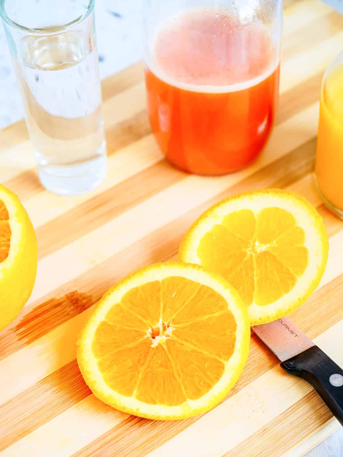 Sliced orange on a wooden board with a knife, a glass of clear liquid, a jar of red liquid, and a glass of orange juice.