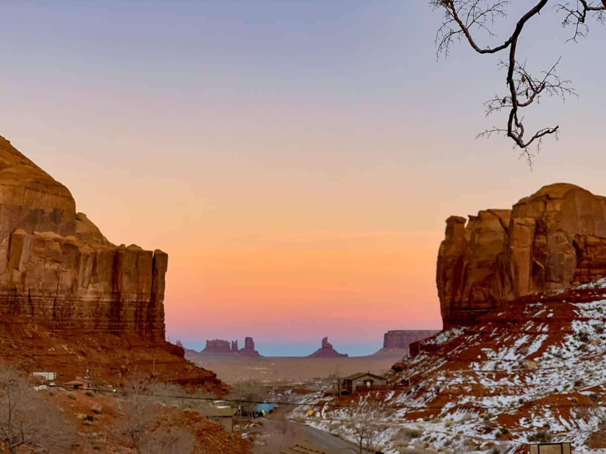 A scenic view of Monument Valley at sunset, featuring large rock formations, a gradient colorful sky, and some snow on the ground. A few small buildings and a road are visible in the foreground.