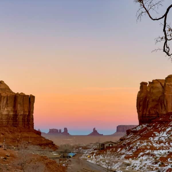 A scenic view of Monument Valley at sunset, featuring large rock formations, a gradient colorful sky, and some snow on the ground. A few small buildings and a road are visible in the foreground.