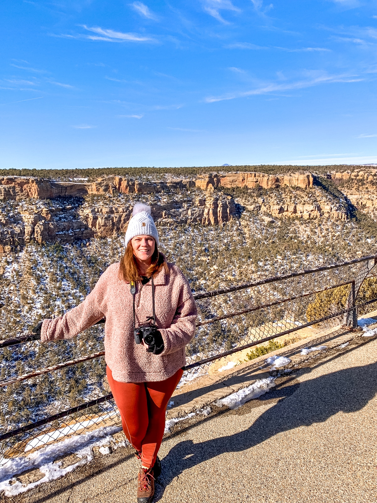 A person in a pink jacket and white beanie standing on a viewpoint overlooking a rocky, snowy canyon, holding a camera. The sky is clear and blue.