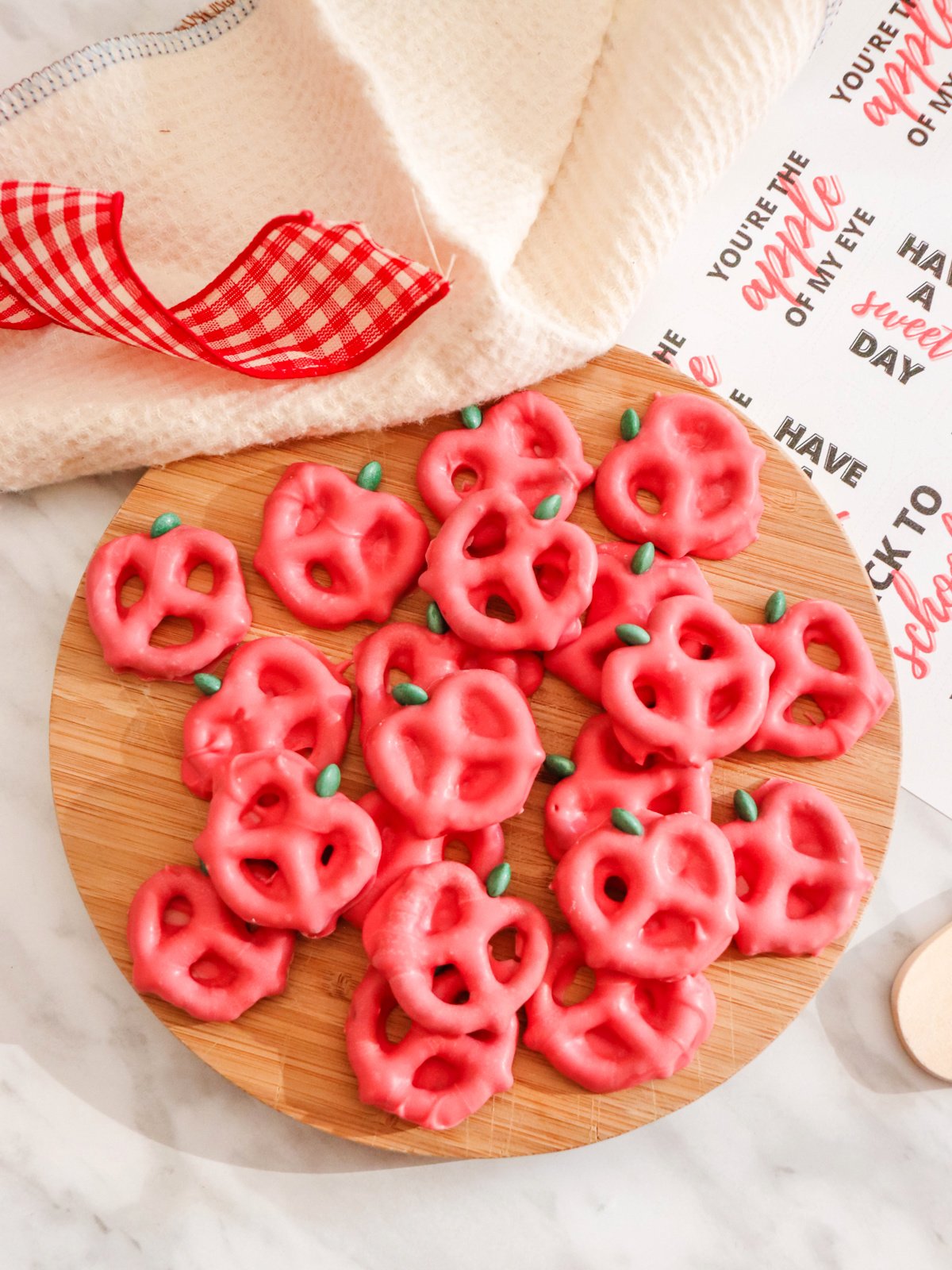 A round wooden board filled with red pretzels shaped like apples, with green leaf decorations on a white surface. A red gingham ribbon and a beige towel are draped in the background.