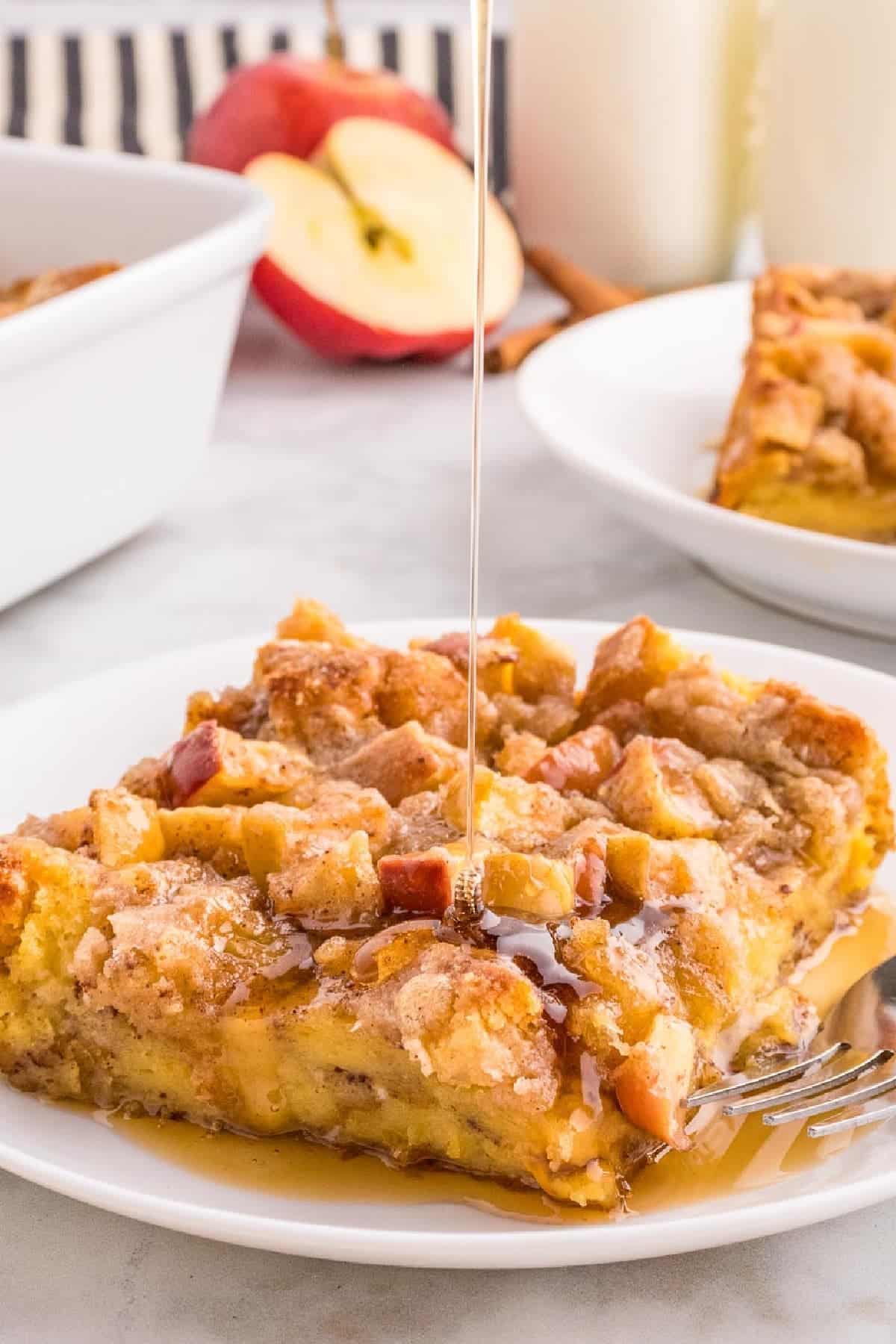 A slice of apple crumble cake on a white plate is being drizzled with syrup. In the background, there is a halved apple and two glasses of milk.