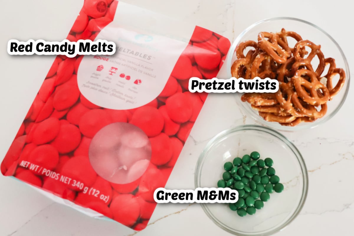 A bag of red candy melts, a bowl of pretzel twists, and a bowl of green M&Ms are arranged on a white surface.