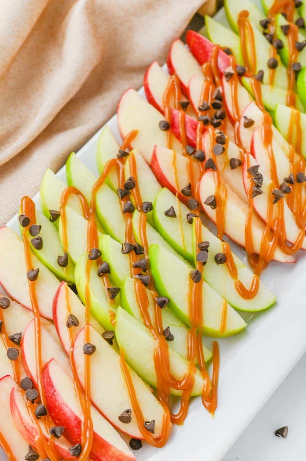 A white platter holds sliced green and red apples, drizzled with caramel sauce and sprinkled with chocolate chips, set against a beige cloth background.