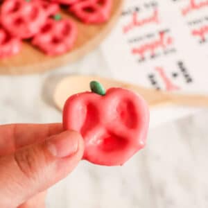 Close-up of a hand holding a pink pretzel resembling an apple with green icing at the top. A wooden spoon and similar pretzels are visible in the blurred background.