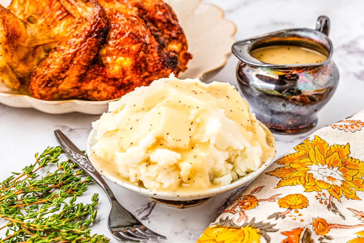 A bowl of mashed potatoes topped with gravy is in the foreground. A roasted chicken and a gravy boat are in the background, with fresh herbs and a floral napkin placed nearby.