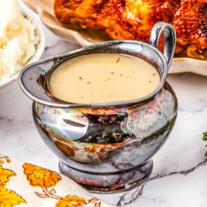 A silver gravy boat filled with light brown gravy sits on a marble countertop. Mashed potatoes and a roasted chicken are visible in the background.