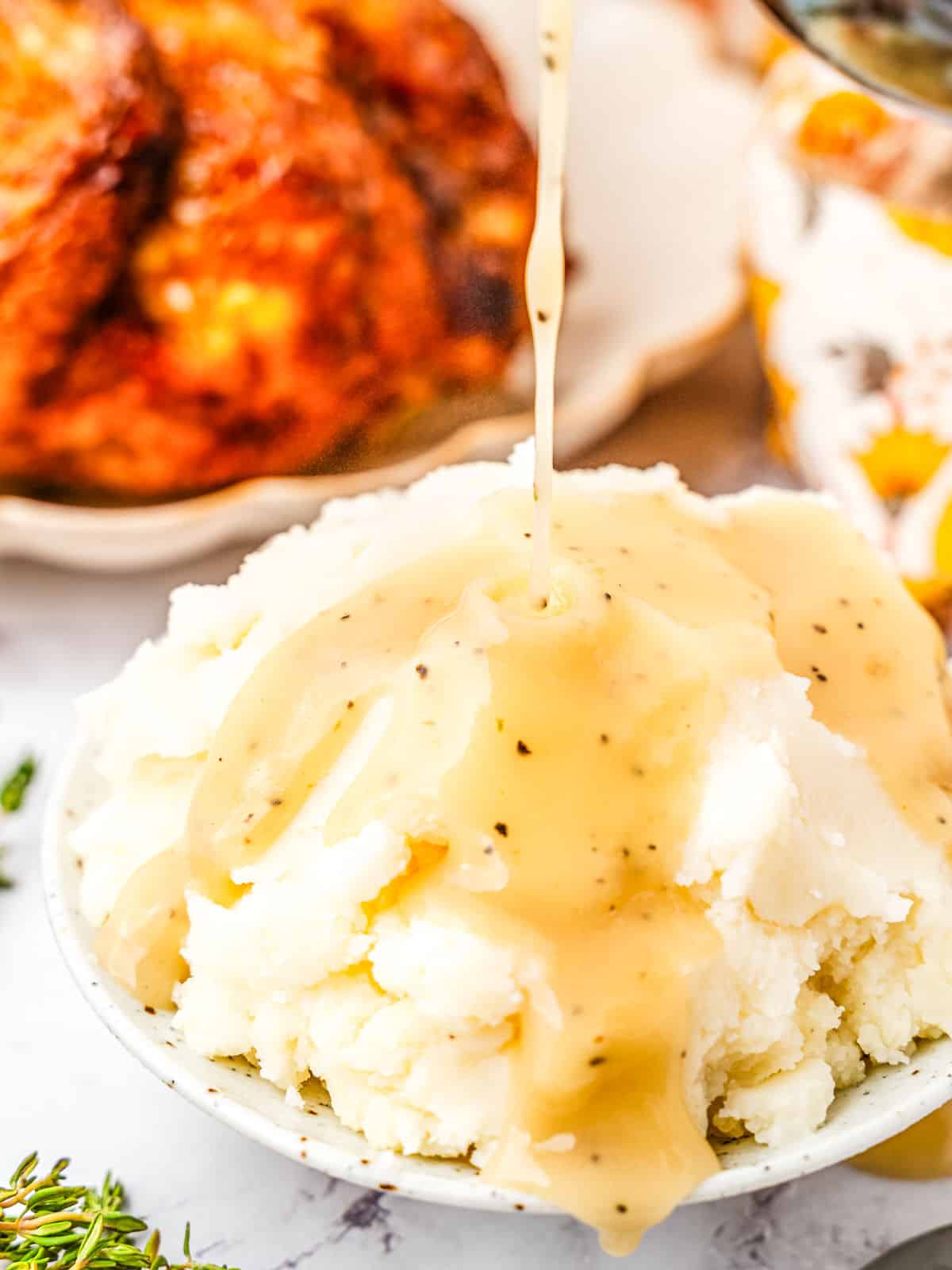 A bowl of mashed potatoes is being drizzled with gravy. In the background, there is a plate of roasted meat.